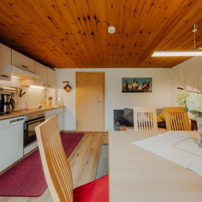 Helle Wohnküche der Ferienwohnung Mittagspitze im Haus Wilma, im gemütlichen, natürlichen Stil mit Holzdecke mit Blick von Esstisch auf Küchenzeile links und Sofa rechts hinten
