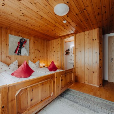 zweites Schlafzimmer der Ferienwohnung Mittagspitze mit Holzdecke und Wand, Blick auf Doppelbett und kleines Bad rechts neben dem Bett