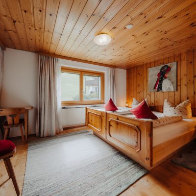 zweites Schlafzimmer der Ferienwohnung Mittagspitze mit Holzdecke im Haus Wilma, Doppelbett neben dem Fenster vor Holzwand und kleinem Tisch in der Zimmerecke